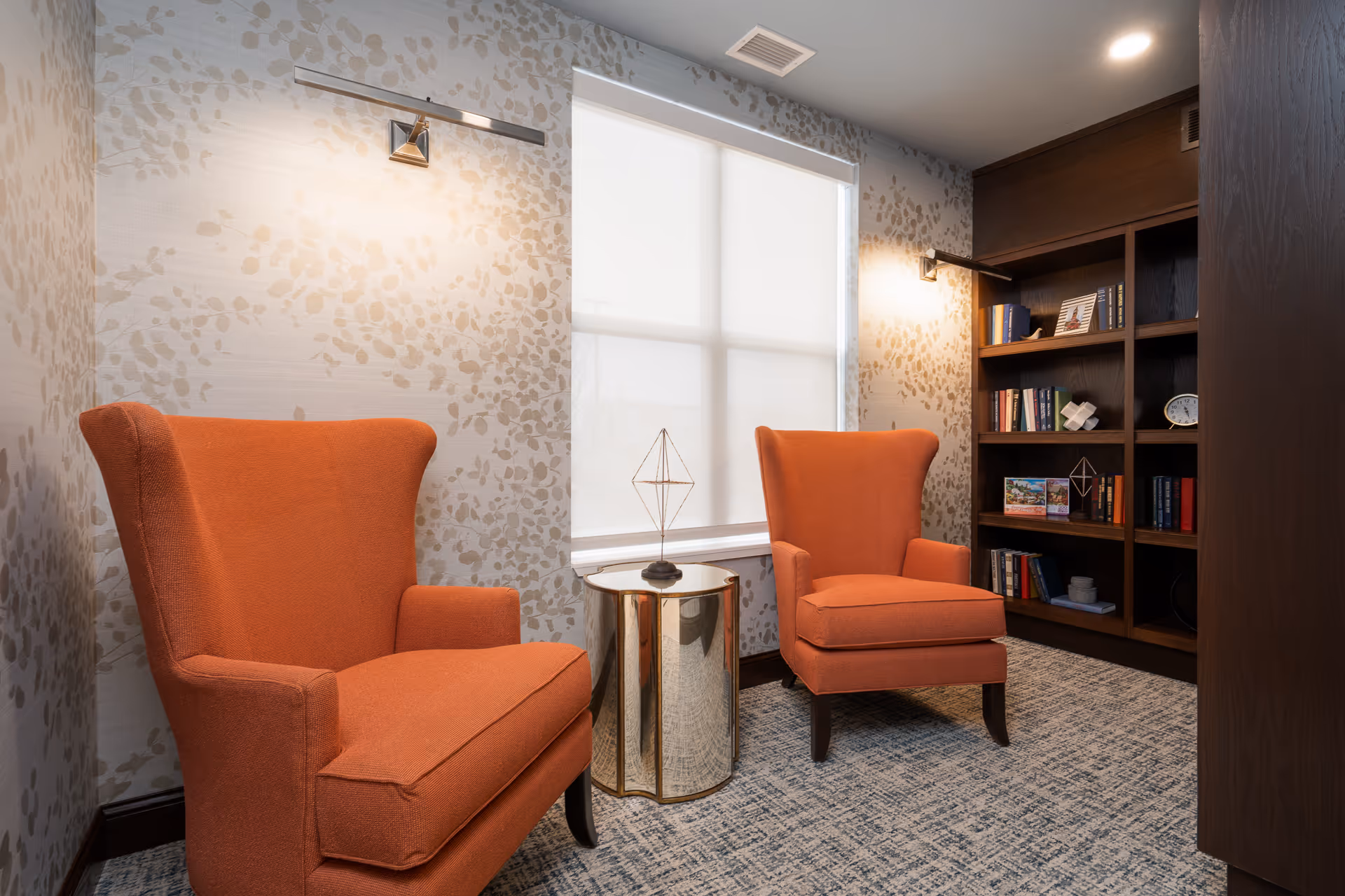 A cozy sitting area with two orange upholstered armchairs facing each other, separated by a small round metallic side table. Behind the chairs is a window with a white shade, and to the right is a wooden bookshelf filled with books and decorative items. The walls have a light-colored wallpaper with a subtle leaf pattern, and the floor is carpeted.