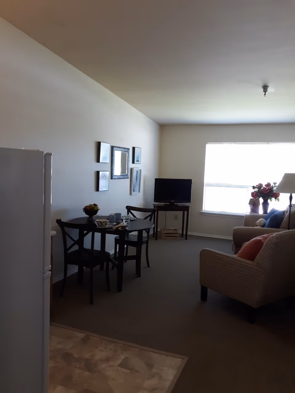 Interior view of a living area in an assisted living facility showing a small dining table with two chairs, a television on a stand, a sofa, an armchair, a window with blinds, and a vase with flowers on a side table.