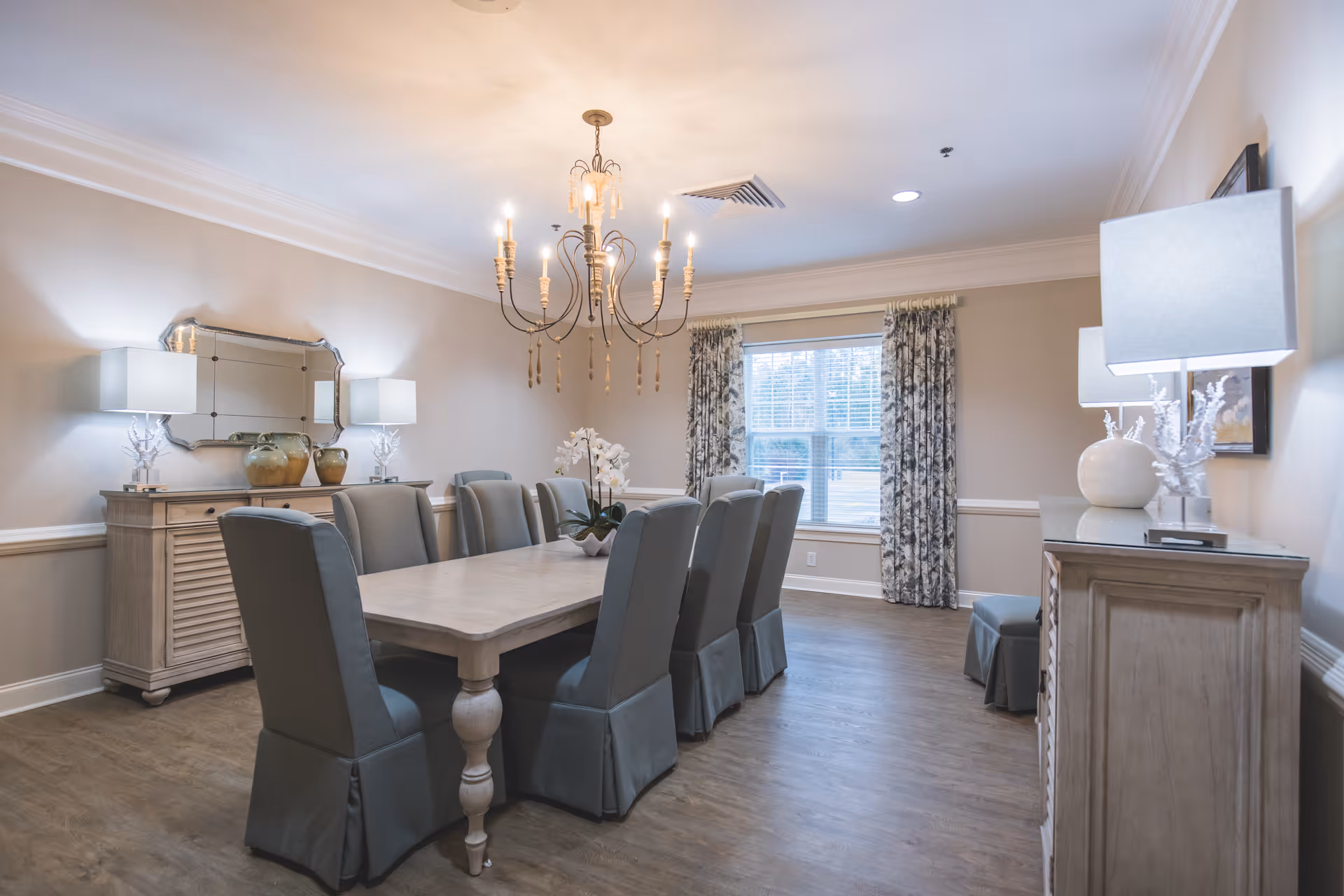 A well-lit dining room with a long rectangular table surrounded by eight cushioned chairs covered in gray fabric. The room features a chandelier hanging from the ceiling, two sideboards with table lamps and decorative items, a large window with patterned curtains, and light-colored walls with white trim.