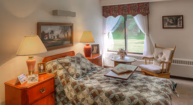 A cozy bedroom in a senior living facility featuring a bed with a patterned quilt, two bedside tables each with a lamp, framed pictures on the walls, a window with sheer white curtains and a valance, a small round table with a flower arrangement, and a cushioned wooden rocking chair.