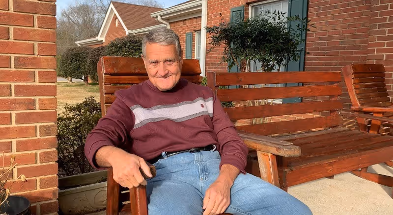 An elderly man wearing a maroon and gray striped sweater and blue jeans is sitting on a wooden bench outside a brick building. There are more wooden benches and some greenery visible in the background.