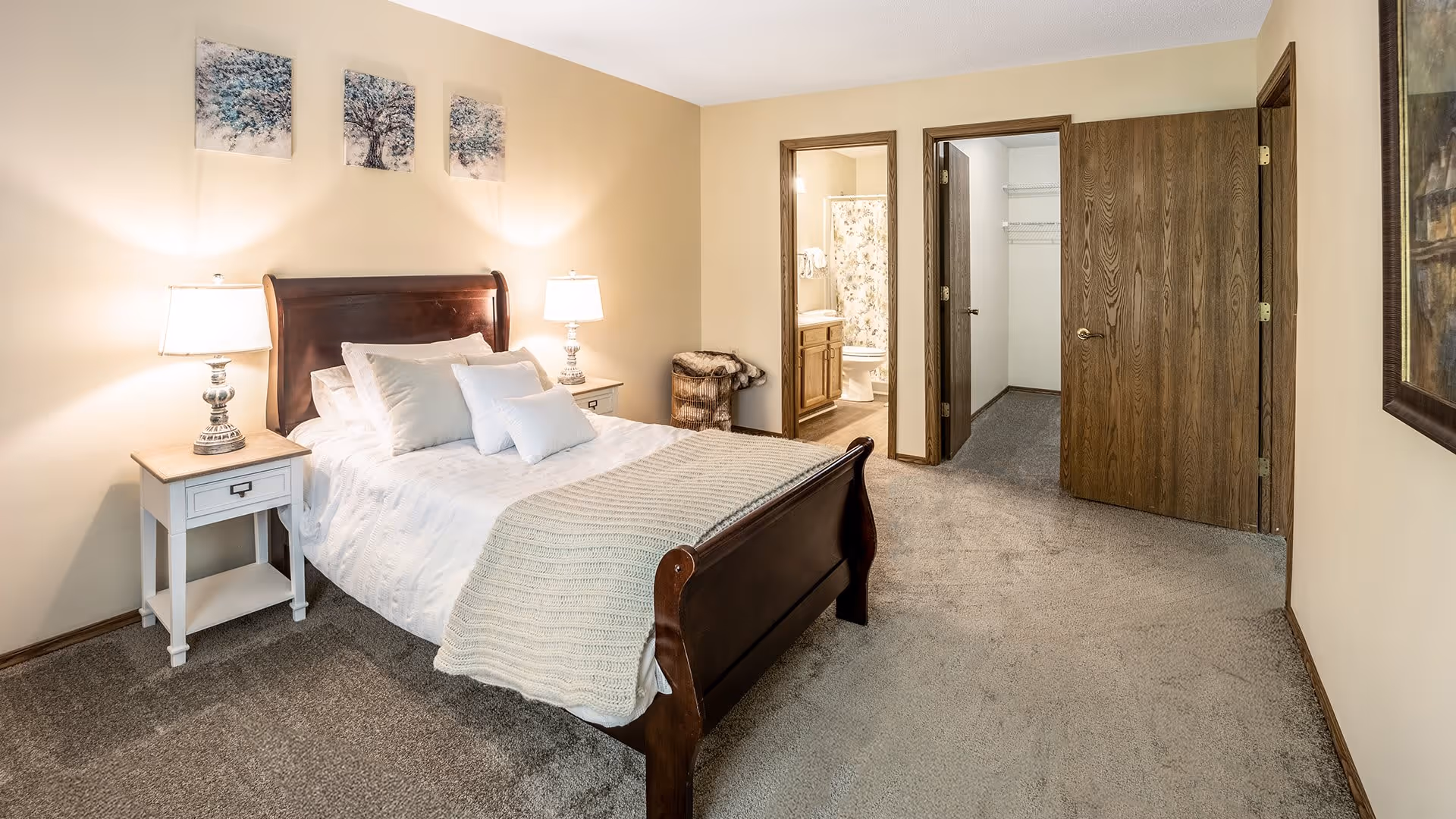 A cozy bedroom with a wooden bed frame, white bedding, and multiple pillows. There are two bedside tables with lamps on each side of the bed. Three pieces of artwork hang above the bed. The room has beige walls and carpeted floors. Two open doors reveal a bathroom with a shower curtain and a walk-in closet.