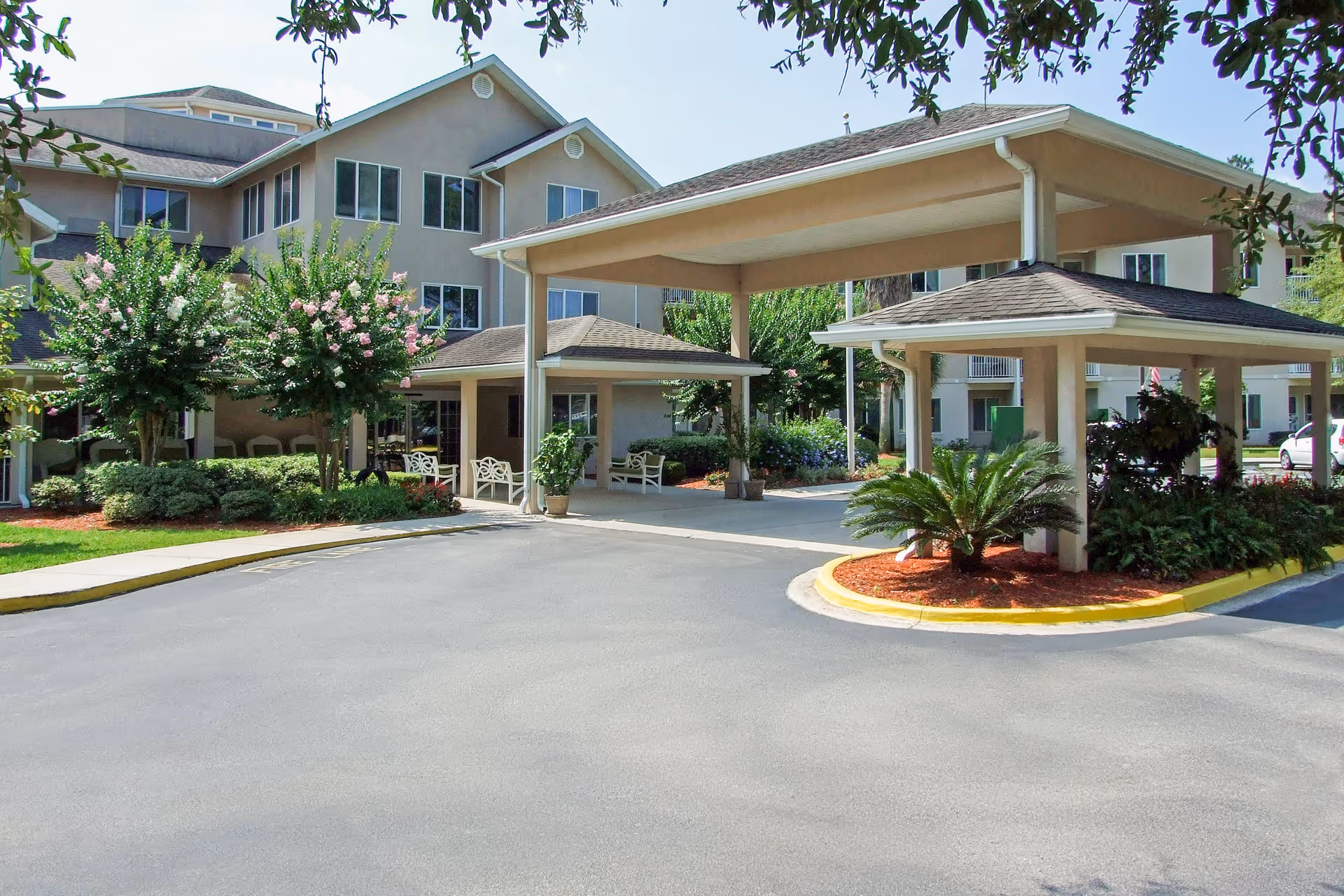 Exterior view of Augustine Landing senior living facility showing a covered entrance with seating areas, landscaped greenery including flowering trees and shrubs, and a multi-story building in the background under a clear sky.