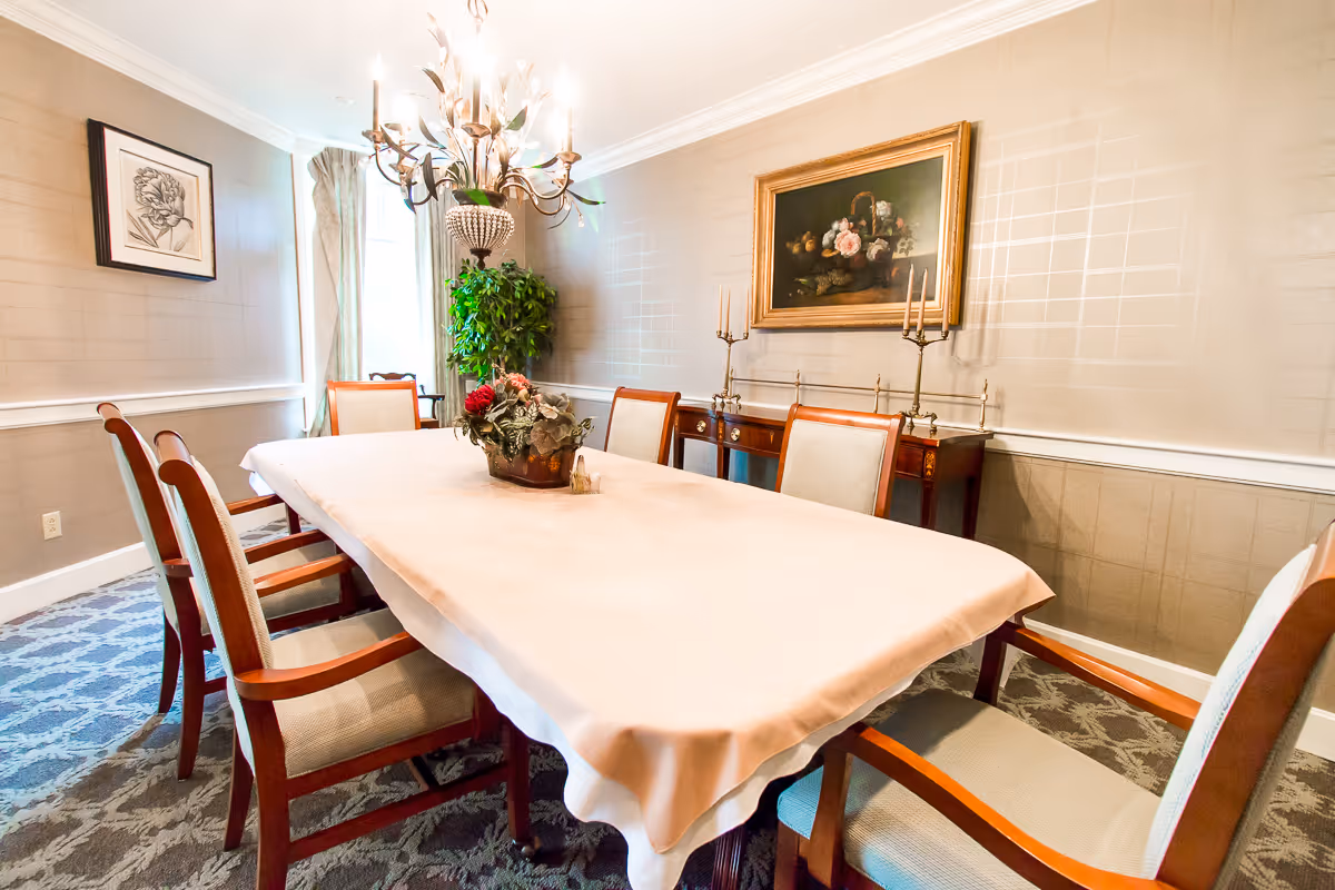 Formal dining room with a long table covered by a pale tablecloth surrounded by wooden chairs, a chandelier overhead, and framed artwork on the walls.