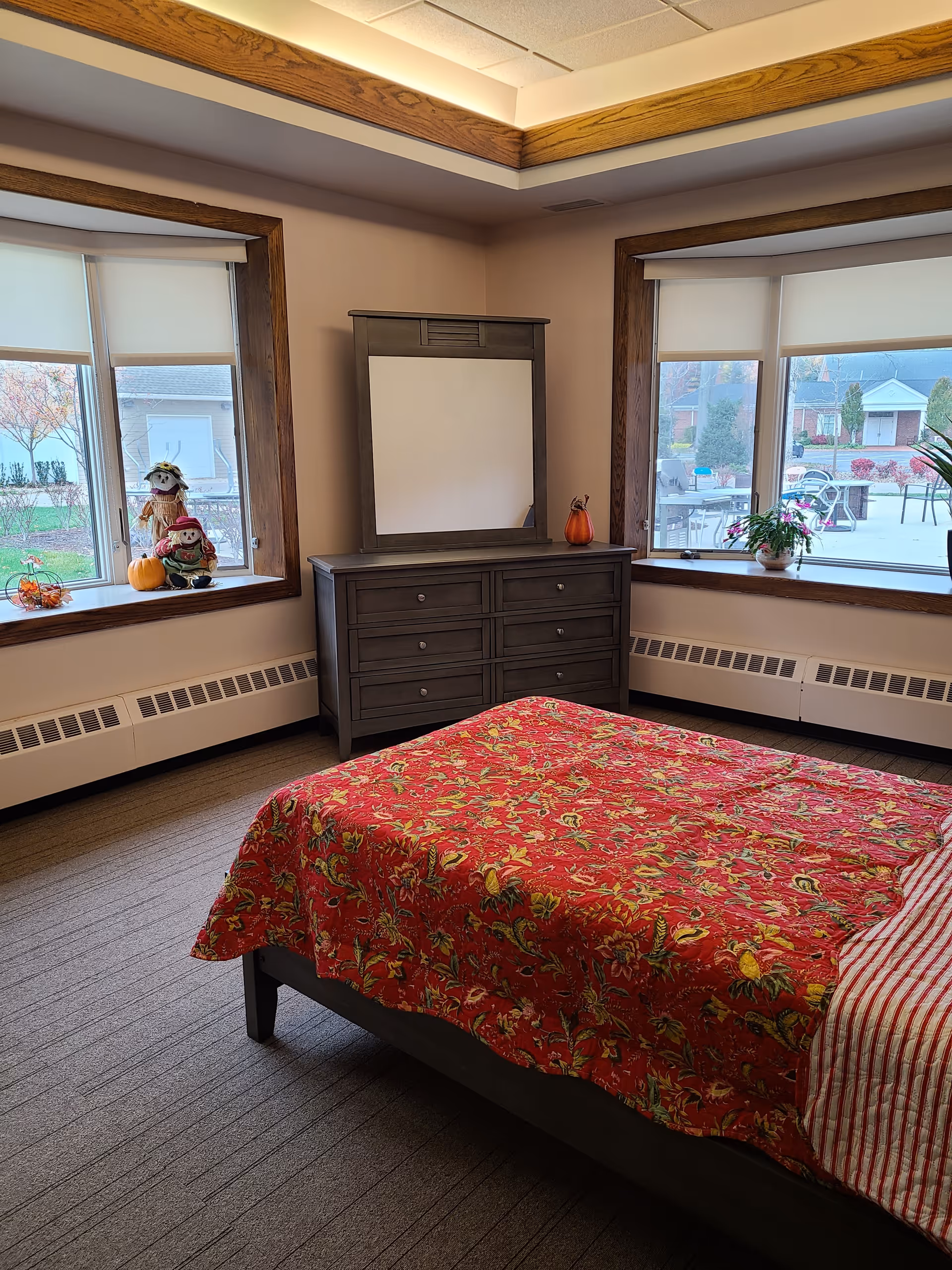 Small bedroom with a bed covered in a red floral quilt, a gray dresser and mirror between two large windows decorated with fall items.