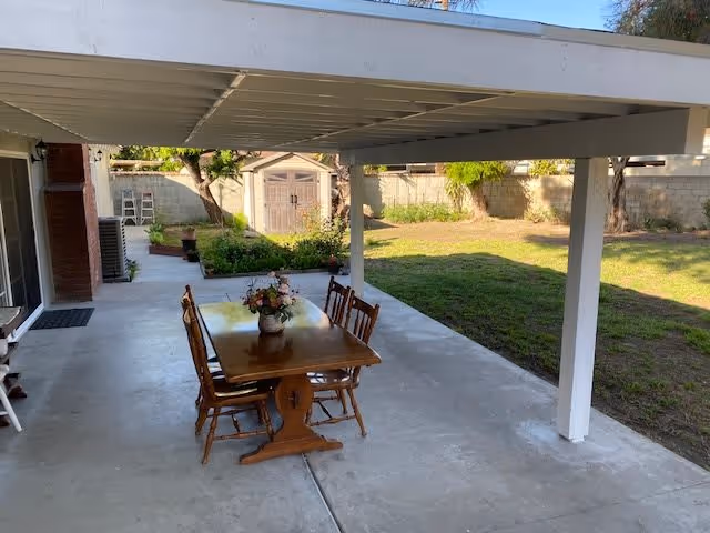 Outdoor covered patio area with a wooden table and four chairs. A flower arrangement is placed on the table. The patio overlooks a grassy backyard with trees and a storage shed against a brick wall fence.