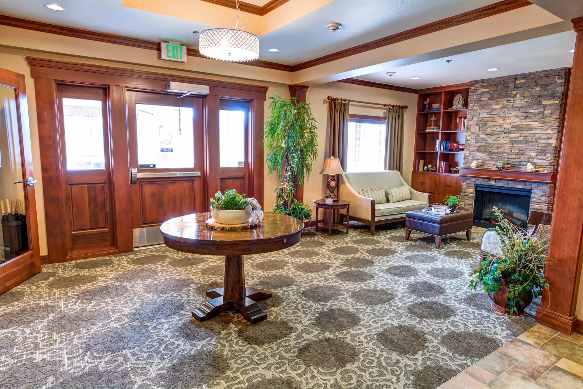 A cozy and well-lit common area with a round wooden table in the center holding a decorative plant. To the right, there is a seating area with a beige loveseat, a brown ottoman, a side table with a lamp, and built-in wooden shelves next to a stone fireplace. Large wooden double doors with glass panels are at the back, and a tall green plant stands near the window with curtains.