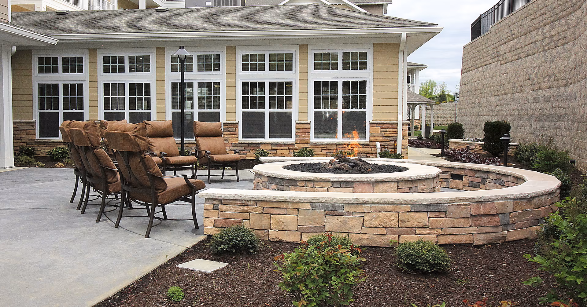 Outdoor patio area at Ivy Creek Gracious Retirement Living featuring a circular stone fire pit with a small fire burning, surrounded by cushioned brown chairs. The patio is adjacent to a building with multiple large windows and beige siding, and there are small bushes and plants around the patio area.