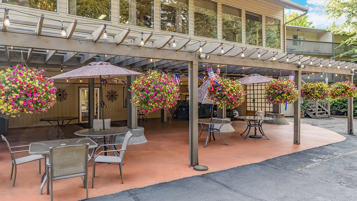 Covered outdoor patio with tables, chairs, umbrellas, hanging flower baskets, and string lights.