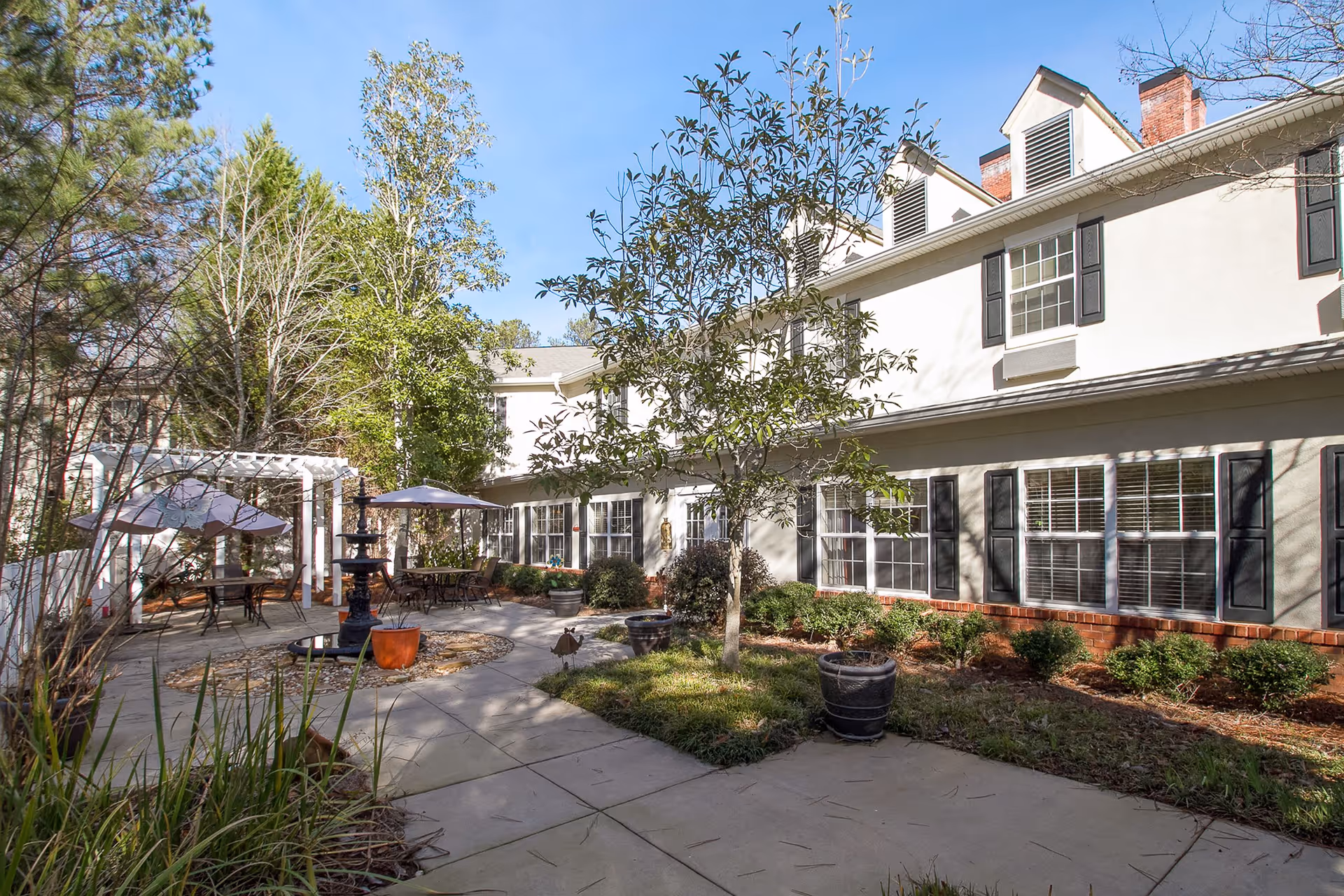 Outdoor courtyard area of a senior living facility with a paved walkway, small trees, shrubs, and patio tables with umbrellas. The building has white walls, black shutters, and multiple windows. A white pergola is visible on the left side.