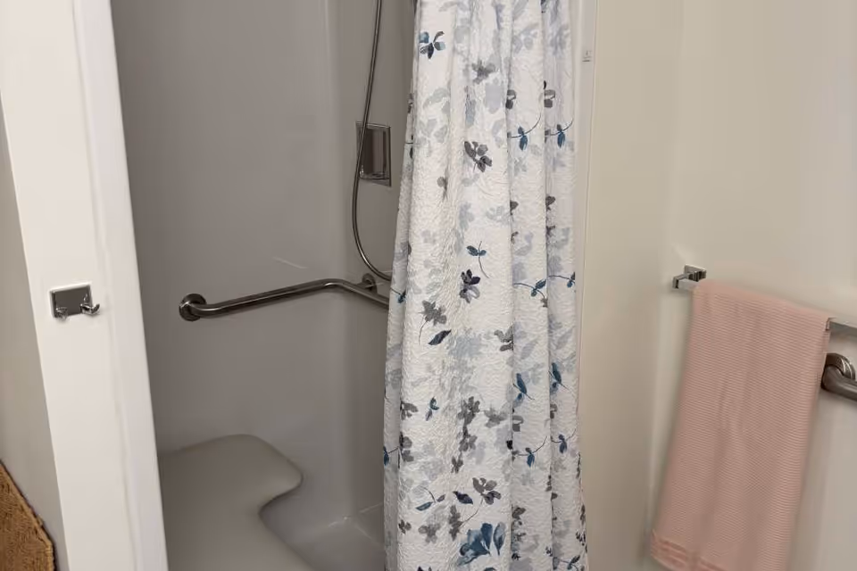 A bathroom shower area with a white and blue floral shower curtain partially drawn. Inside the shower, there is a built-in seat and a horizontal grab bar. A pink towel hangs on a towel bar outside the shower on the right wall.