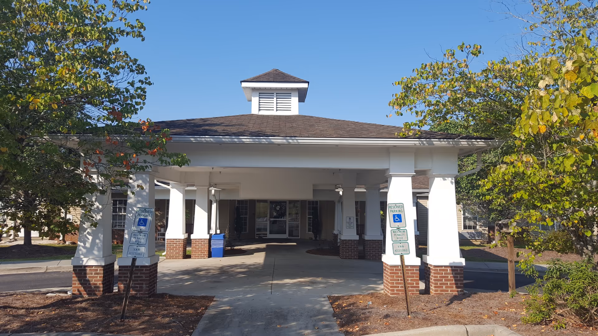 Covered entrance and porte-cochere of a senior living building with white columns, brick bases, accessible parking signs, and surrounding trees.