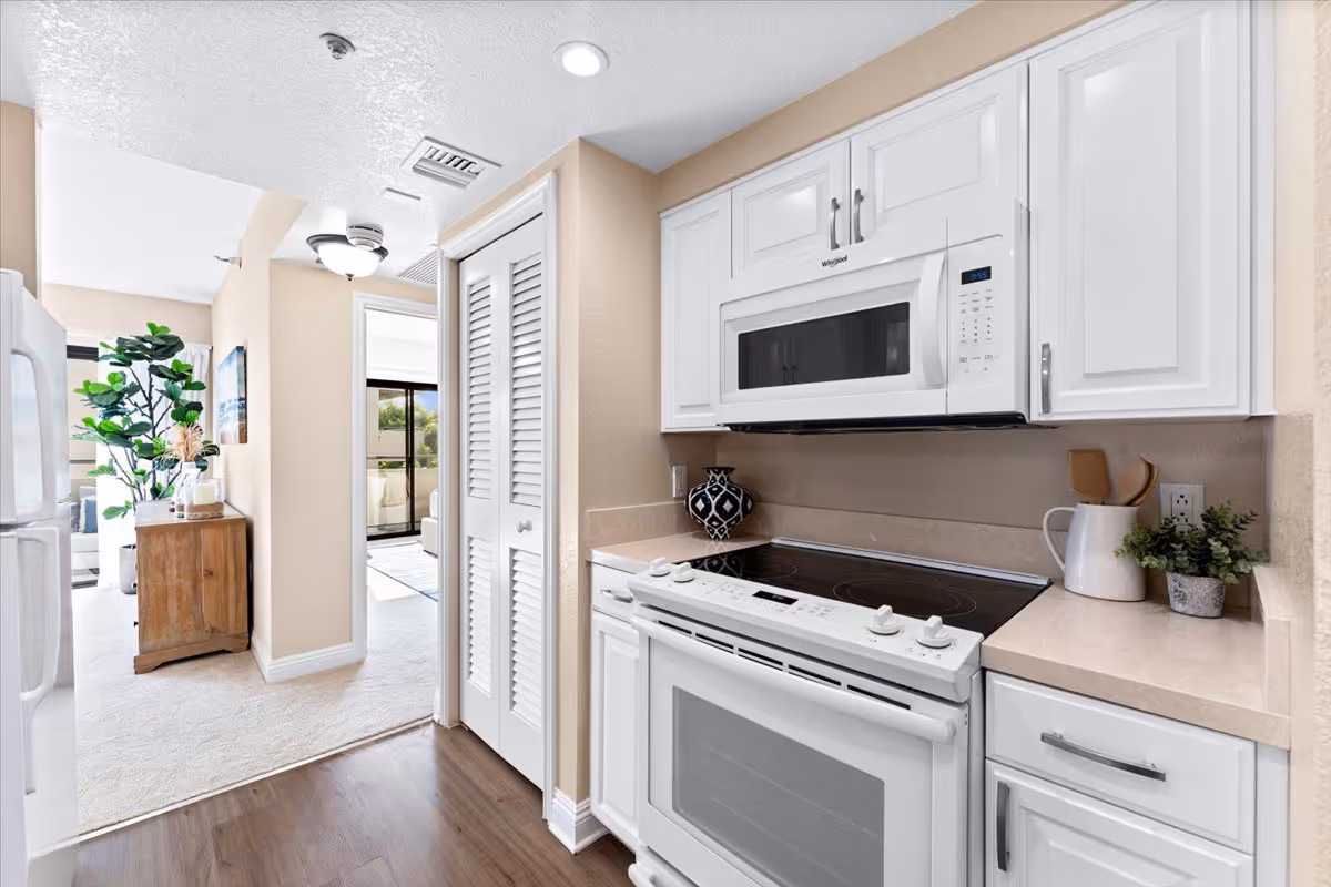 A bright kitchen area with white cabinets, a white electric stove, and a white microwave above it. The countertops are beige, and there are decorative items including a black and white vase and a white pitcher with wooden utensils. The kitchen opens into a hallway leading to a living area with a wooden cabinet, a large green plant, and a window letting in natural light.