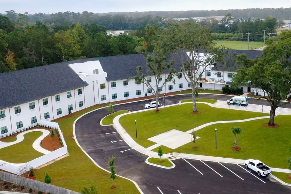 Aerial view of a two-story white assisted living building with a curved driveway, landscaped lawn, and a few parked vehicles.
