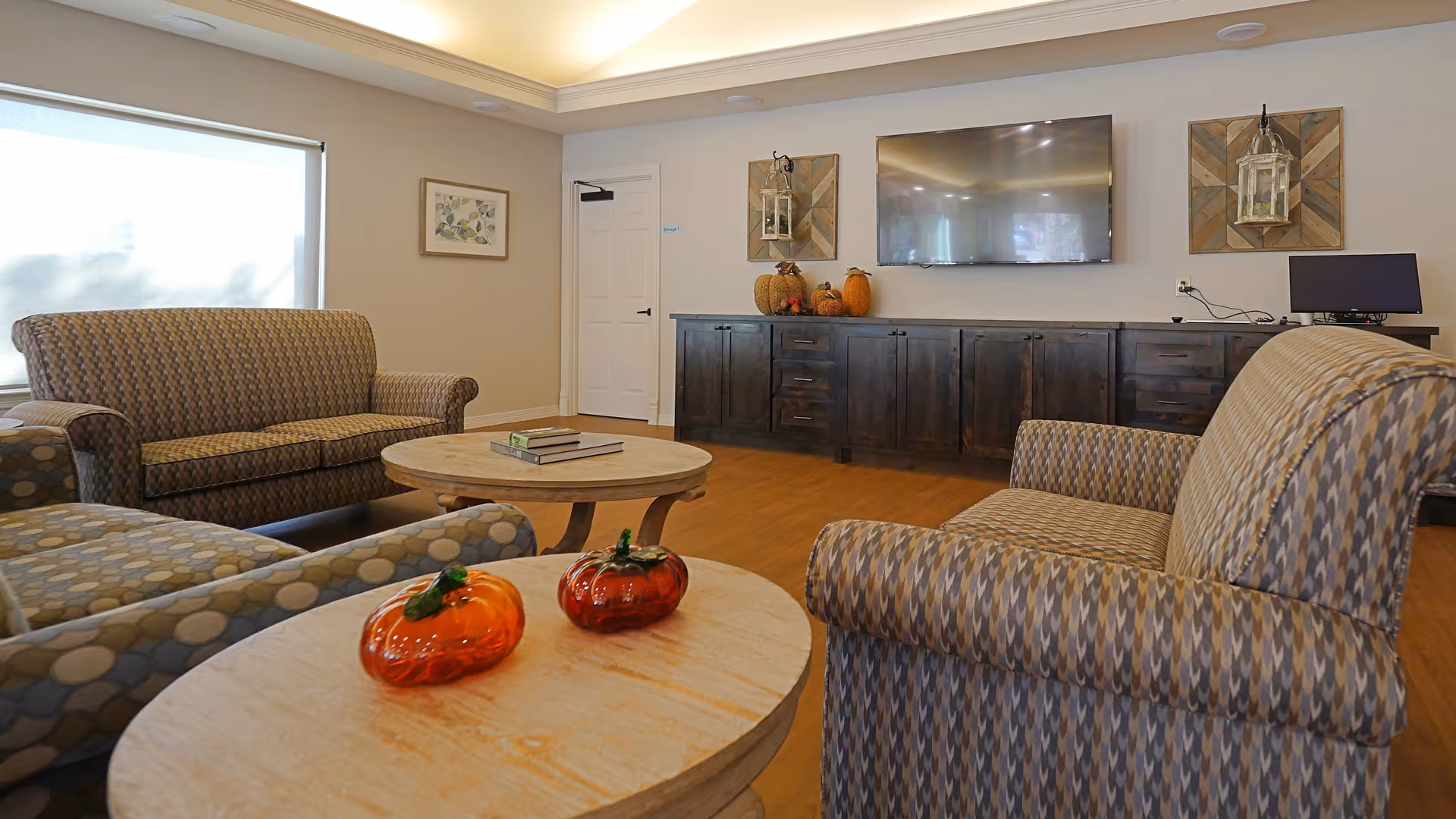 A cozy living room area with patterned armchairs and a sofa arranged around two wooden coffee tables. On the tables are two decorative glass pumpkins. The room features a large flat-screen TV mounted on the wall above a long dark wooden cabinet, with fall-themed decorations including pumpkins. The walls are light-colored, and there is a window with a shade partially drawn, a framed picture, and a white door in the background.