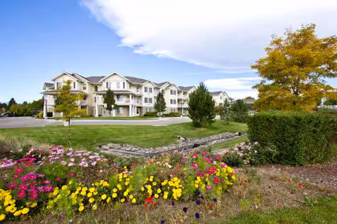 A large, multi-story residential building with balconies surrounded by well-maintained green lawns, colorful flower beds, and trees under a partly cloudy sky.