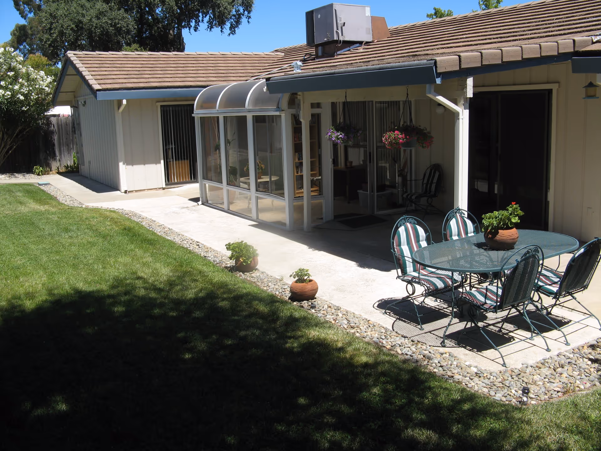 Backyard patio with a lawn, outdoor dining table and chairs beside a sunroom attached to a single-story building.