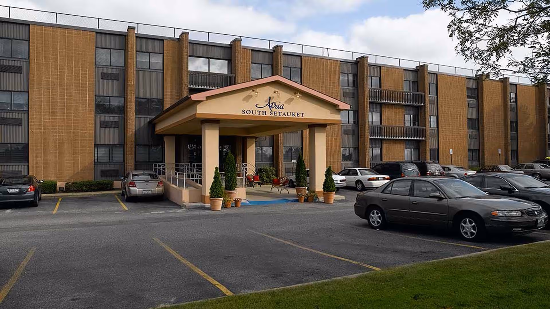 Exterior view of a three-story senior living facility building with a covered entrance labeled 'Atria South Setauket'. Several cars are parked in the parking lot in front of the building, and there are potted plants near the entrance.