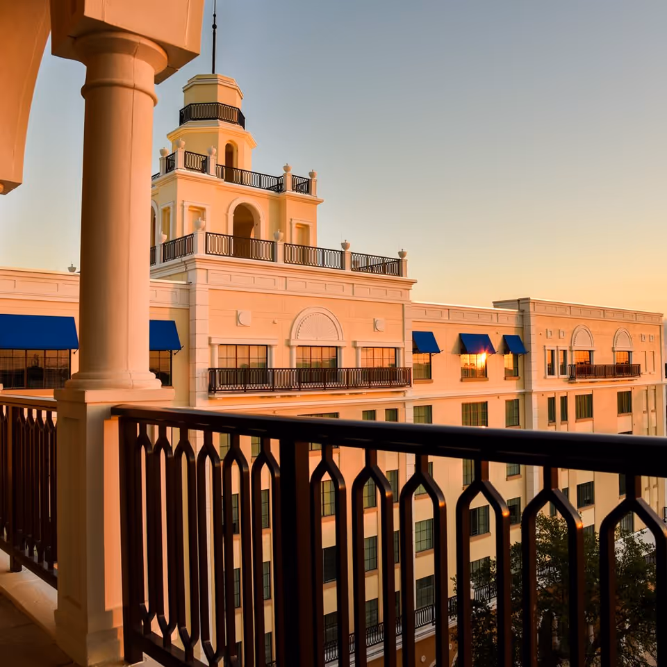 View of a multi-story building with classical architectural design featuring a tower with balconies and blue awnings over windows, seen from a balcony with decorative black railing during sunset.