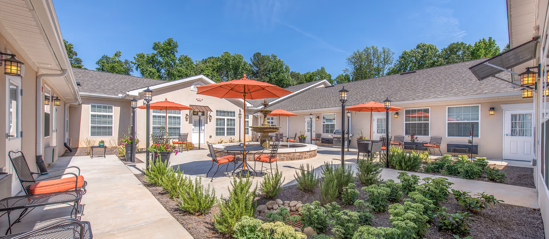 Sunny courtyard with patio tables, orange umbrellas, a central fountain, and surrounding single-story buildings.