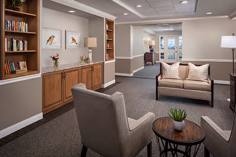 A cozy senior living facility common area with two upholstered armchairs and a small round wooden table with a potted plant in the foreground. A loveseat with two pillows is positioned against the wall. Built-in wooden shelves with books and decorative items flank a countertop with two framed bird prints and a table lamp. The space has neutral-colored walls, carpeted floors, and recessed ceiling lights, with a hallway and double doors visible in the background.