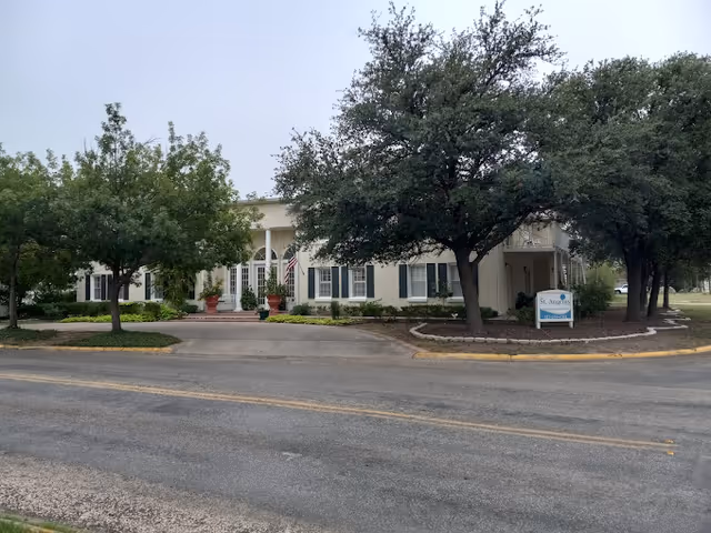 Front exterior view of a single-story building with white walls and dark shutters, surrounded by trees and landscaping. An American flag is displayed near the entrance, and a sign reading 'St. Angelus' is visible near the driveway.
