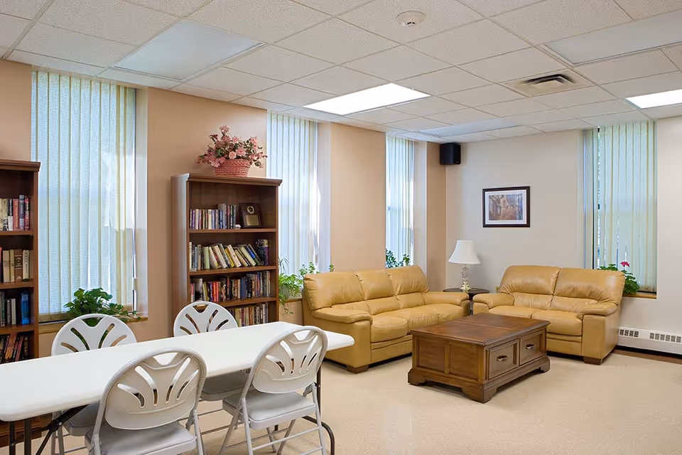A cozy common area with two tan leather sofas arranged around a wooden coffee table. There are tall windows with vertical blinds letting in natural light. A white table with four white chairs is in the foreground. Bookshelves filled with books and a basket of flowers on top are against the wall. A framed picture and a table lamp are also visible.