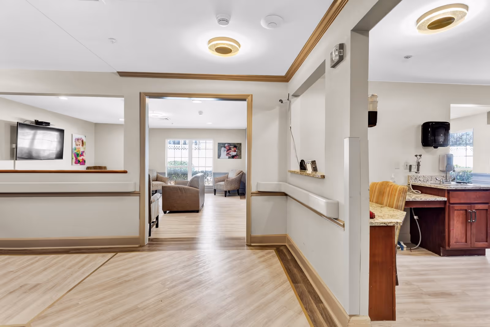 Interior view of a senior living facility showing a hallway leading to a common sitting area with armchairs and a large window. To the right, there is a small kitchenette or reception area with a granite countertop, sink, and a striped chair. The walls are light-colored with wood trim, and the floor has a light wood pattern. Ceiling lights are visible, and there are framed pictures on the walls.