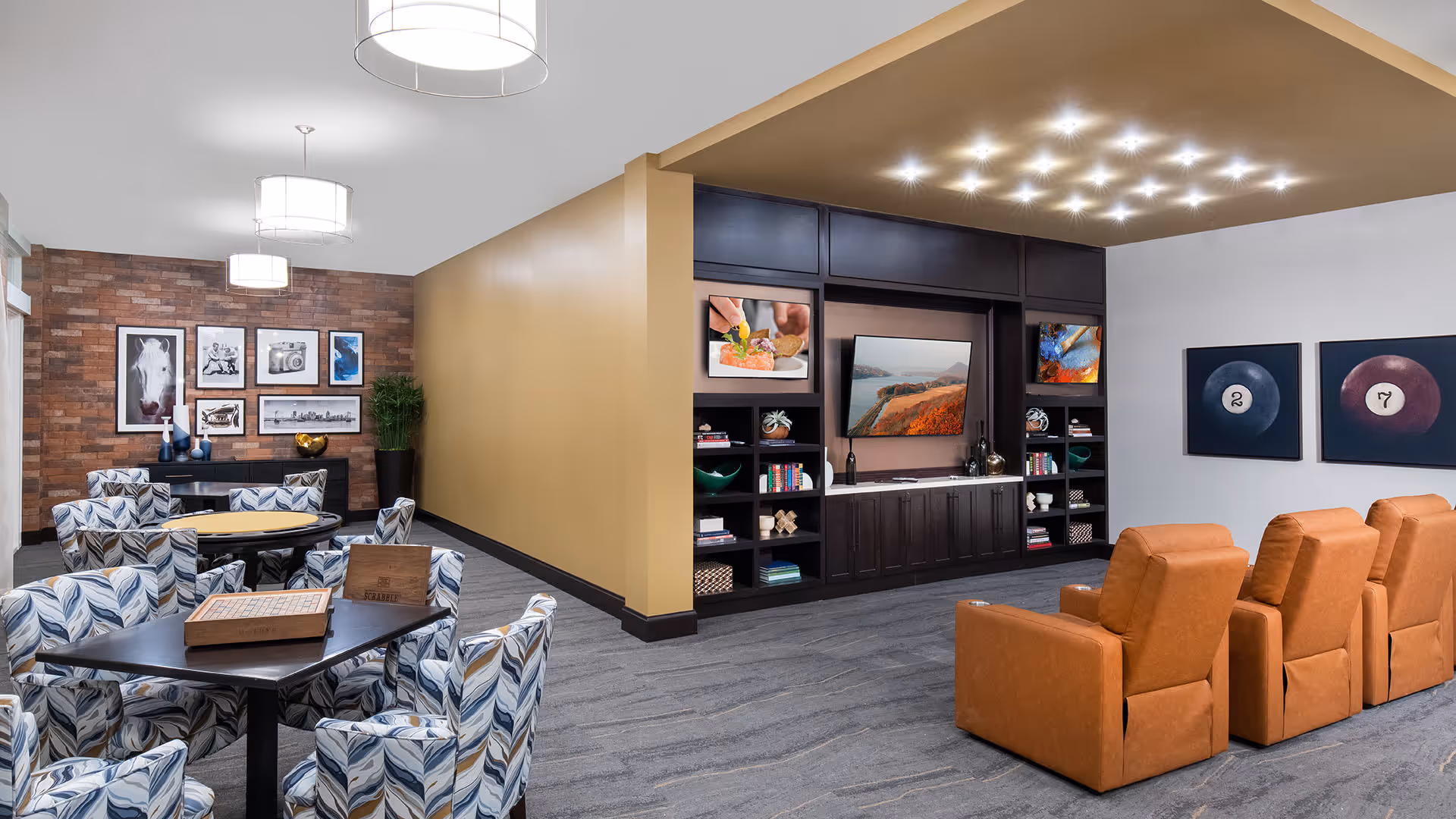 A modern senior living facility common area with a game table surrounded by patterned chairs on the left and a media area with four brown recliners facing a large TV on the right. The walls feature framed artwork and decorative shelving with books and plants. The ceiling has contemporary light fixtures.