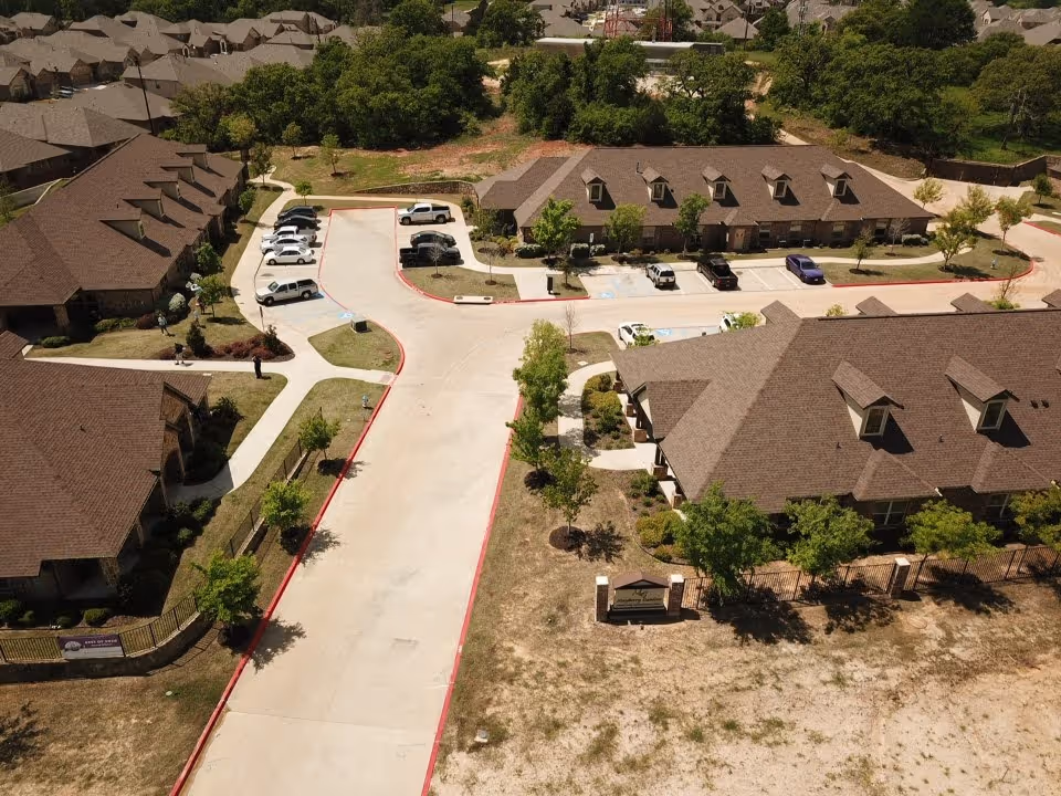 Aerial view of Mayberry Gardens Assisted Living and Memory Care Homes showing multiple brown-roofed buildings, parking areas with cars, sidewalks, trees, and surrounding greenery.