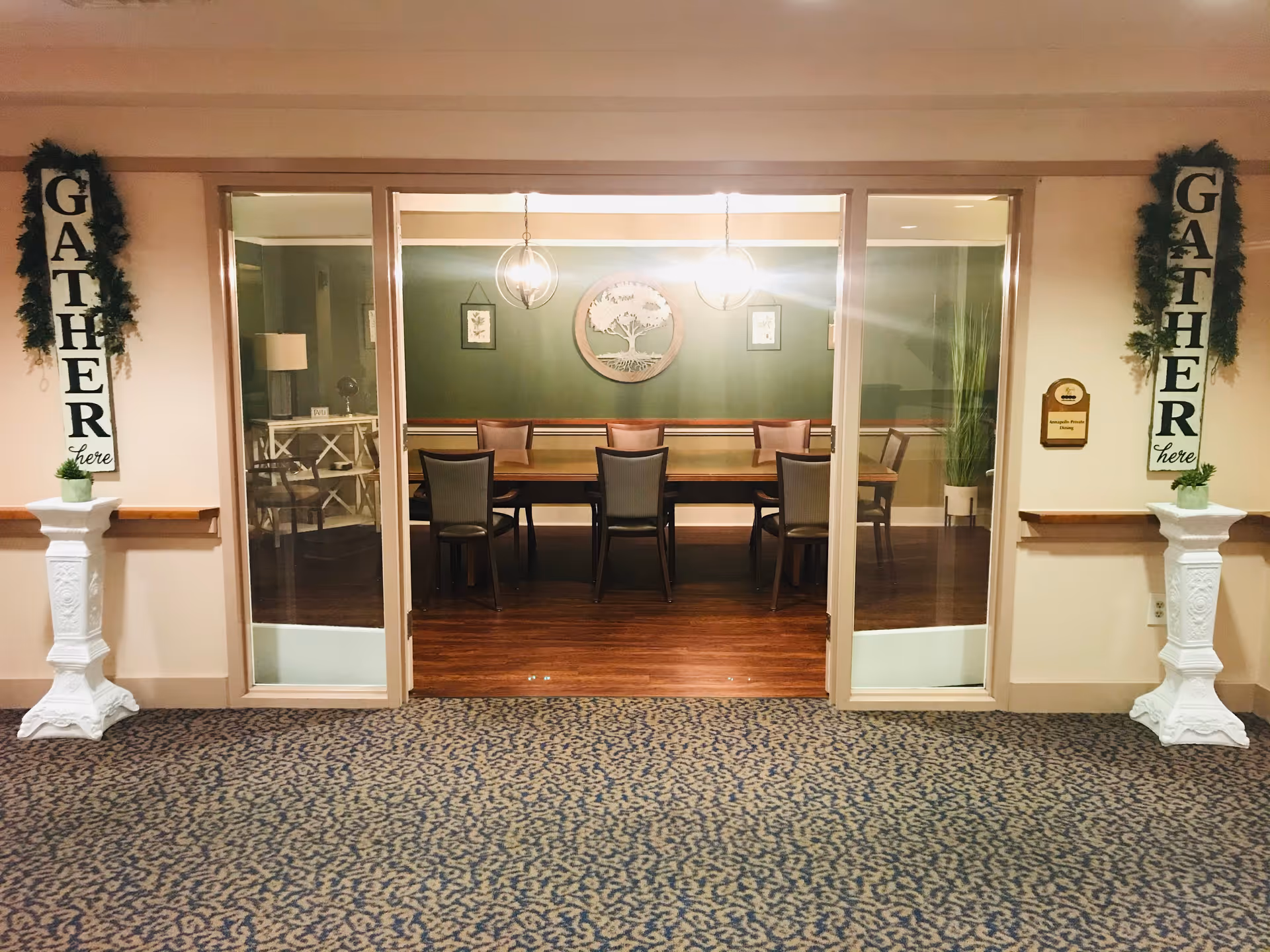 Interior view of a senior living community dining room with a long wooden table and eight chairs. The room has green walls decorated with a large circular tree artwork and two hanging pendant lights. On either side of the entrance are white pedestals with small potted plants and vertical signs that read 'GATHER here' adorned with greenery.