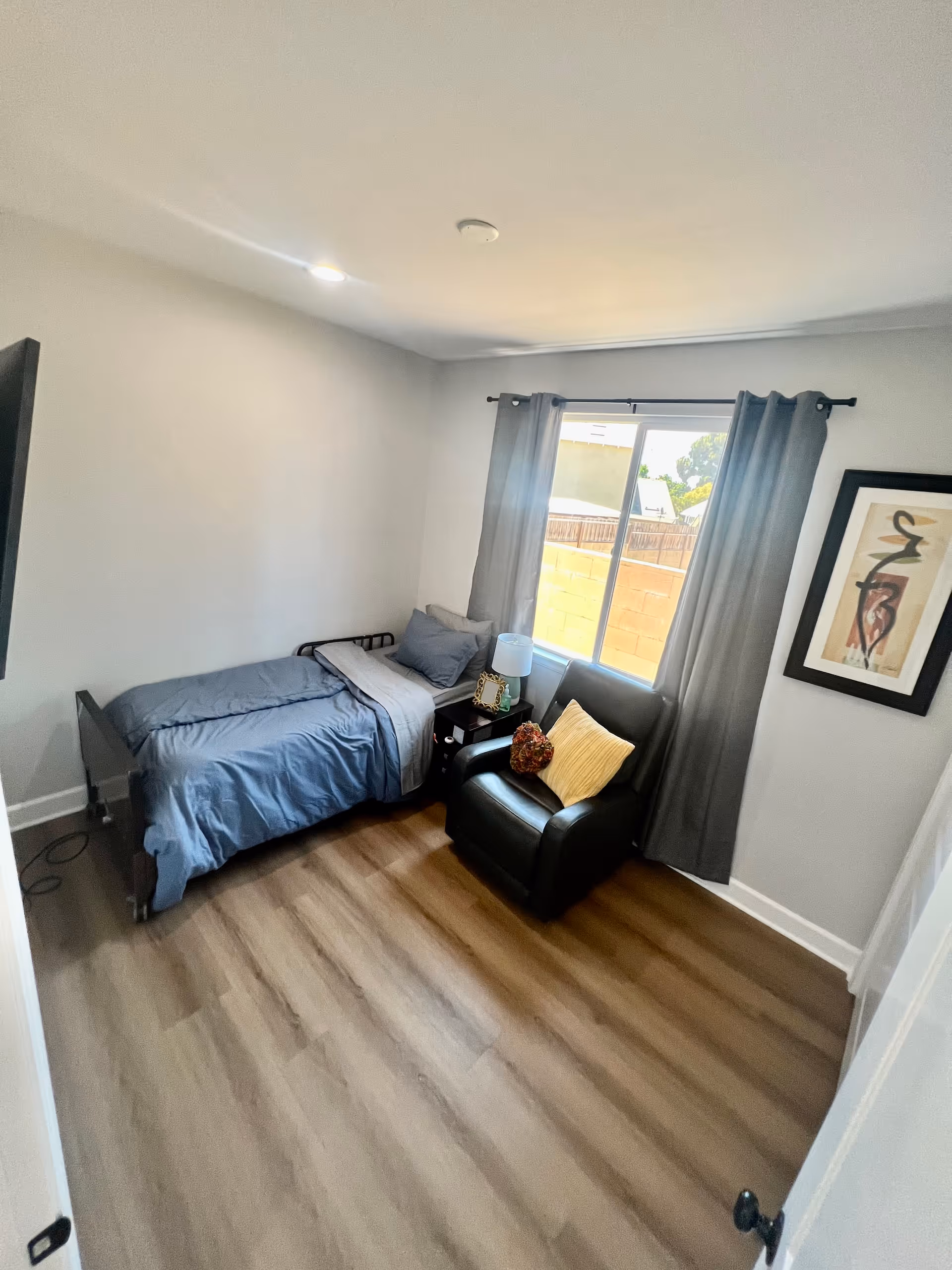 A small bedroom with a single bed covered in blue bedding, a black armchair with a yellow pillow and a decorative cushion, a nightstand with a lamp and picture frame, a window with gray curtains, and a framed abstract artwork on the wall.