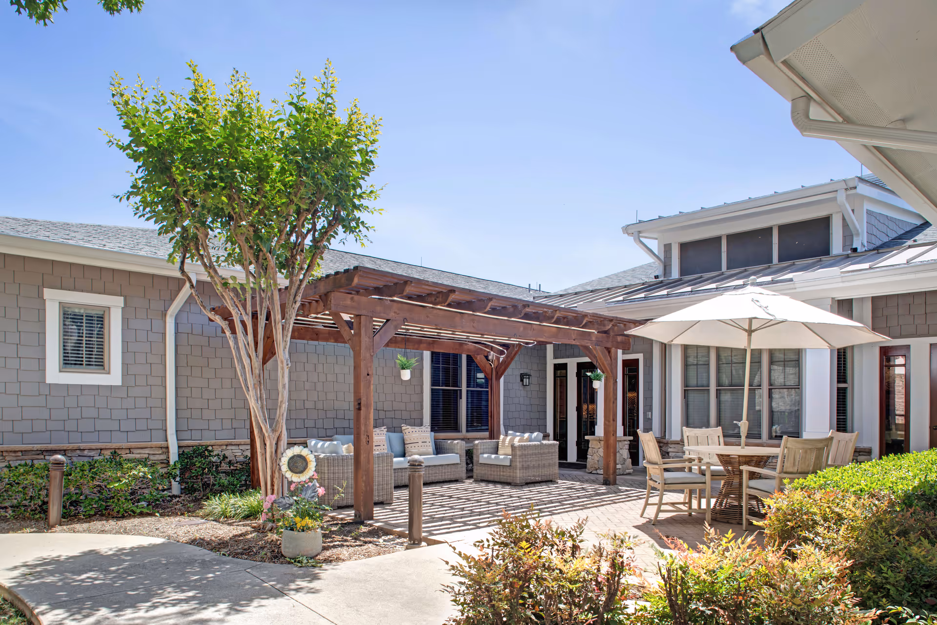 Outdoor patio area at Sunrise of Fort Worth featuring a wooden pergola with hanging plants, cushioned wicker seating, a round table with chairs under a large white umbrella, surrounded by greenery and shrubs under a clear blue sky.