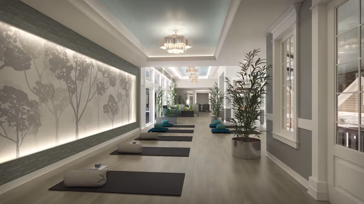 A spacious, well-lit room with yoga mats and bolsters neatly arranged on the floor, surrounded by large potted plants. The walls feature a decorative panel with tree silhouettes backlit by soft lighting. The ceiling has recessed lighting and modern chandeliers, creating a calm and serene atmosphere.