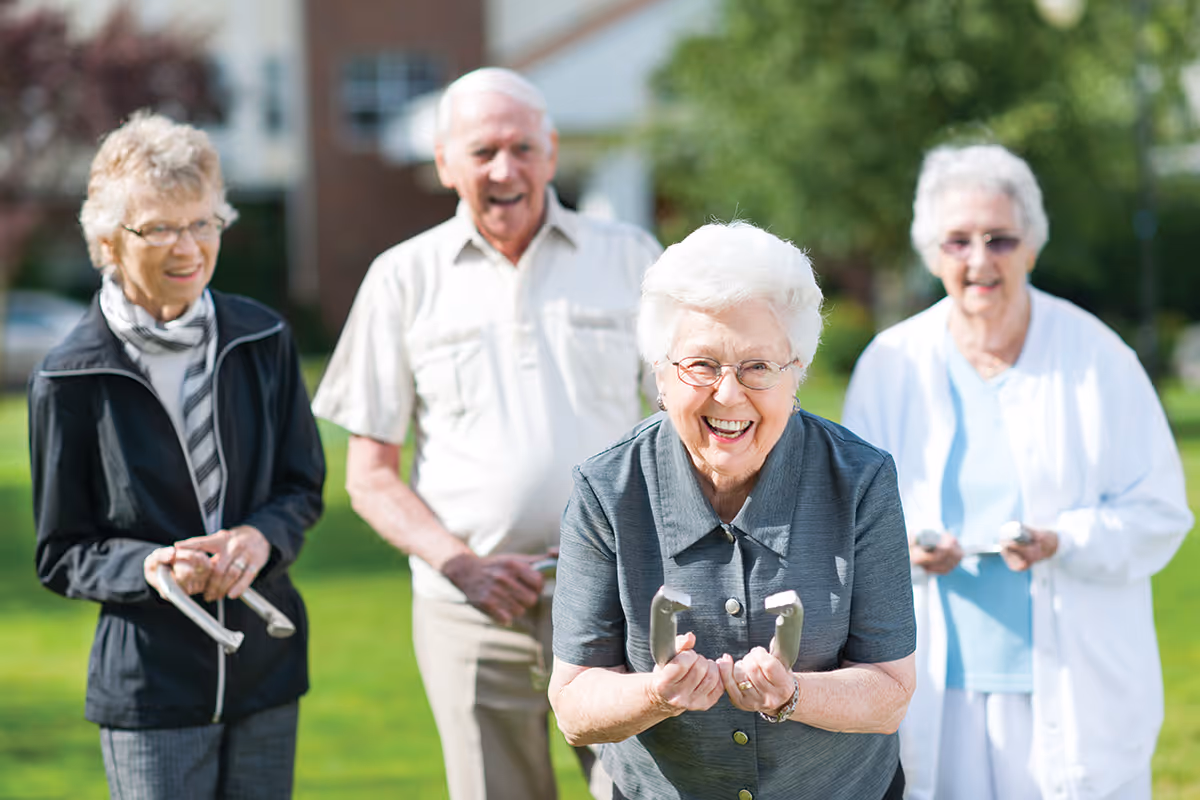 Four elderly people outdoors on a sunny day, smiling and holding horseshoes, appearing to play a game of horseshoes on a green lawn with blurred houses and trees in the background.