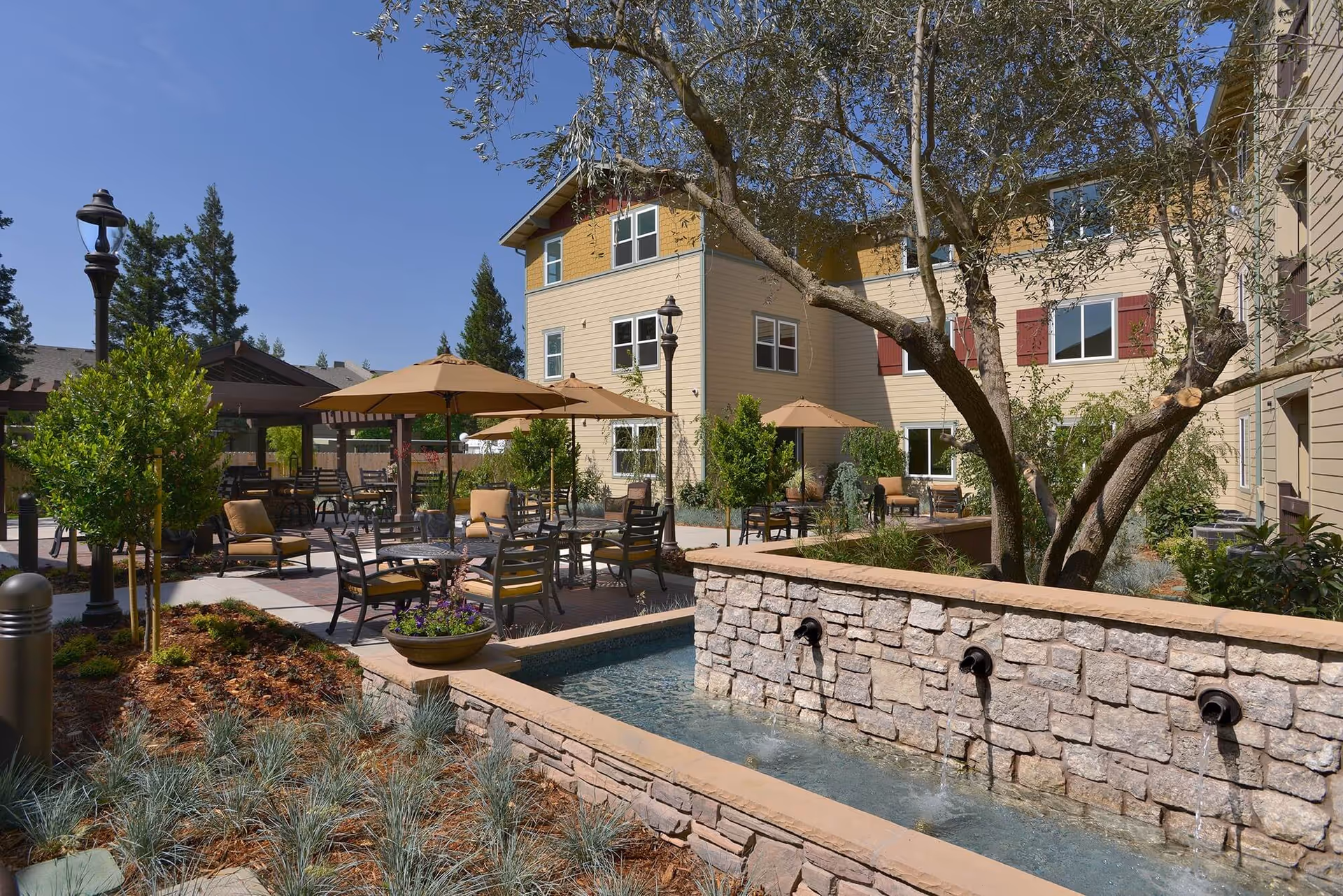 An outdoor courtyard with seating under umbrellas, a stone fountain wall, and beige residential buildings in the background.