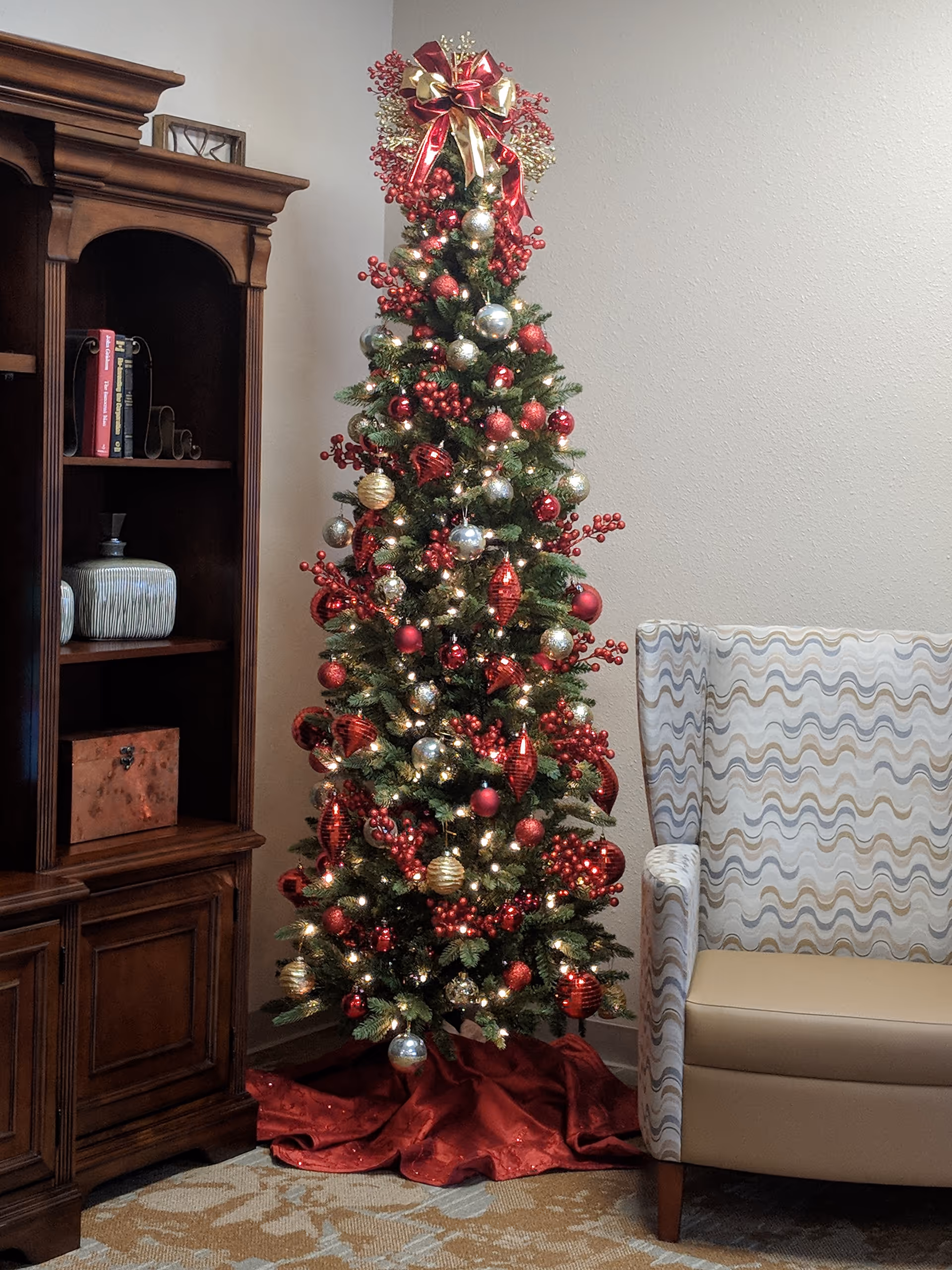 A decorated Christmas tree with red, gold, and silver ornaments and lights stands in a corner next to a wooden cabinet and a patterned armchair in a cozy living room setting.