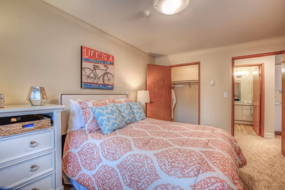 A cozy bedroom with a bed covered in a patterned pink and white comforter and blue decorative pillows. A white nightstand with a basket and a lamp is beside the bed. On the wall above the bed is a framed picture of a bicycle with the text 'LIFE IS A beautiful ride'. The room has beige walls, a closet with an open door, and a doorway leading to a bathroom.