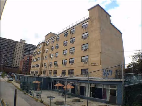 A multi-story beige nursing home with a fenced courtyard and patio tables.