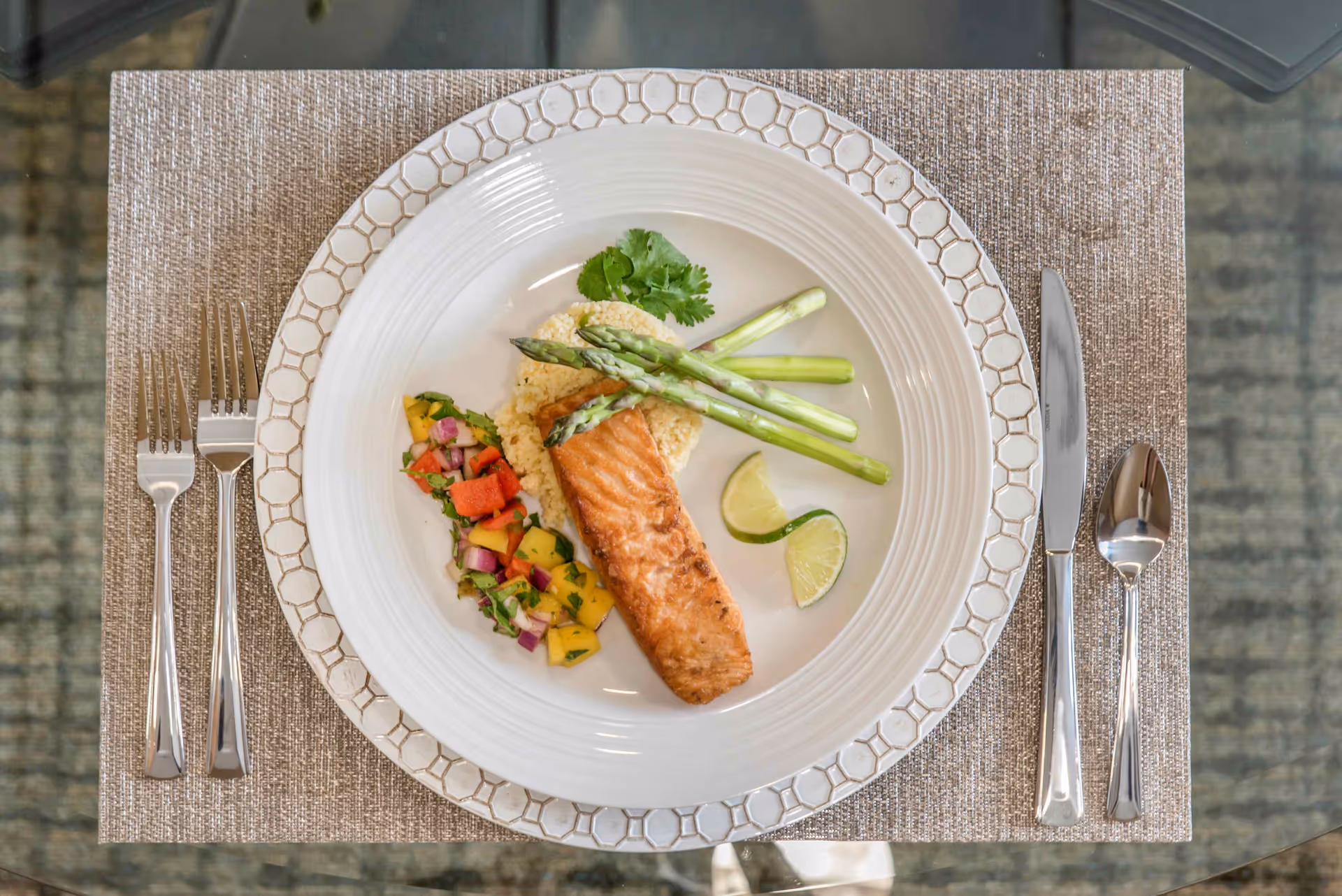 A plated meal consisting of a grilled salmon fillet on a bed of couscous, garnished with three asparagus spears, two lime wedges, and a side of colorful diced vegetables including red onion, mango, and red bell pepper. The plate is set on a beige placemat with a patterned charger underneath, and silverware including two forks on the left and a knife and spoon on the right.