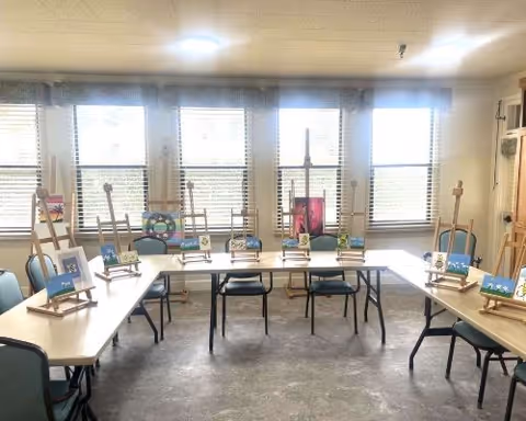 Bright activity room with U-shaped tables and chairs arranged around easels holding small canvases in front of large windows.