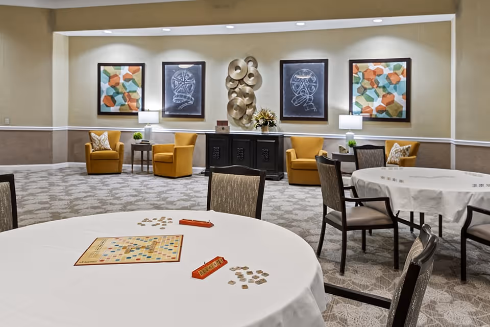 A well-lit common area with round tables covered in white tablecloths and chairs arranged around them. One table has a Scrabble game set up with tiles and racks. In the background, there are four mustard yellow armchairs paired with small side tables and lamps. The walls are decorated with colorful abstract paintings and metallic circular wall art. The carpet has a subtle patterned design.