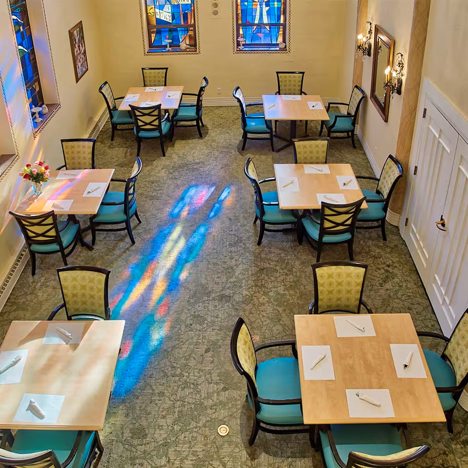 Interior view of a dining room with several square wooden tables, each set with napkins and utensils. The chairs have light green patterned backs and teal seats. Colorful light from stained glass windows reflects on the carpeted floor. The walls are light-colored with framed artwork and wall sconces.