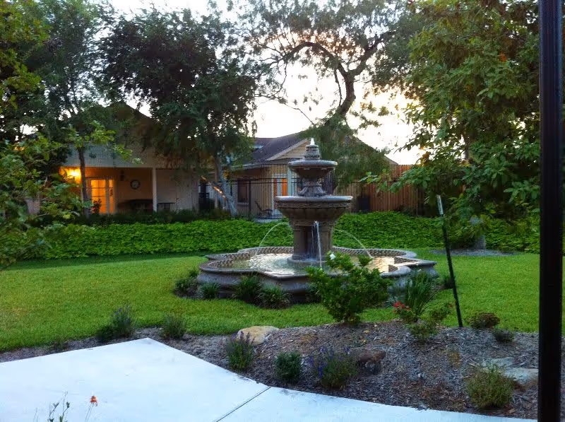A serene outdoor garden area featuring a multi-tiered water fountain surrounded by green grass, plants, and trees. In the background, there is a building with a porch and warm lighting visible through the windows.