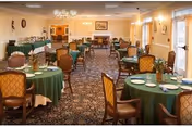 A dining room with multiple round tables covered with green tablecloths, each set with plates, glasses, and silverware. The room has upholstered chairs, a patterned carpet, chandeliers, and warm lighting. There are windows along one side and a fireplace at the far end.