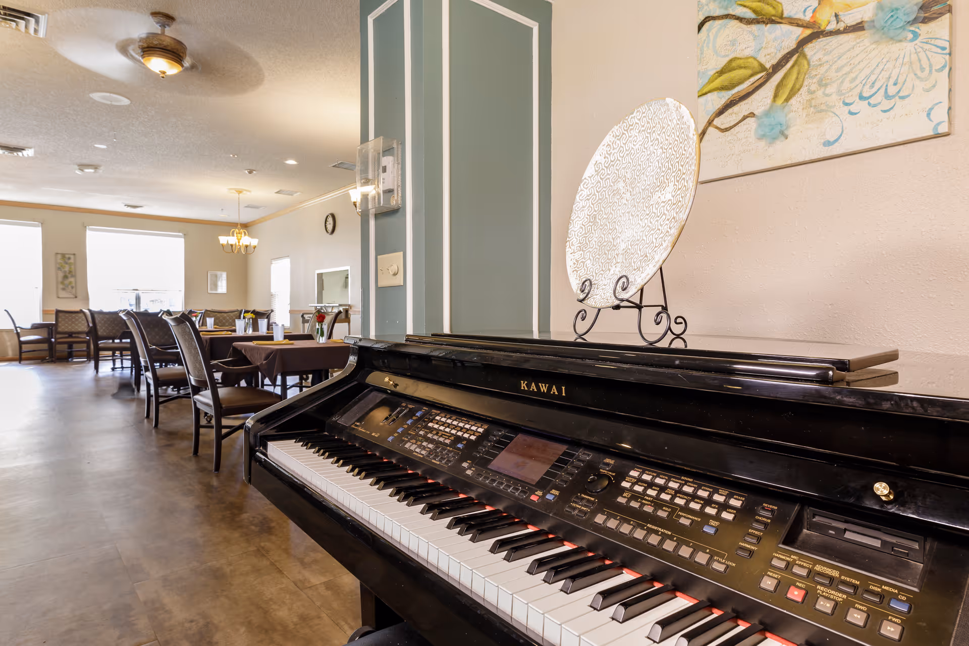 Interior view of a senior living facility dining area with multiple tables and chairs arranged neatly. In the foreground, there is a black Kawai electronic piano with a decorative plate and a floral painting on the wall behind it. The room is well-lit with natural light coming through large windows and ceiling lights.