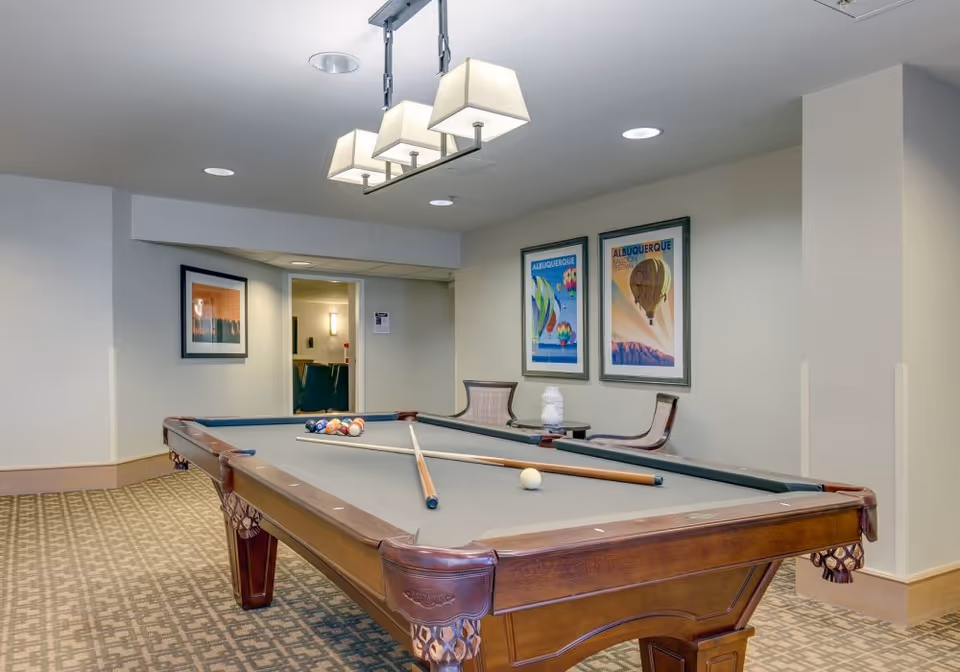 Indoor room with a pool table in the center, two pool cues resting on the table, and a set of billiard balls arranged. The room has patterned carpet flooring, beige walls with framed artwork featuring Albuquerque hot air balloons, two chairs and a small table against the wall, and a modern ceiling light fixture above the pool table.