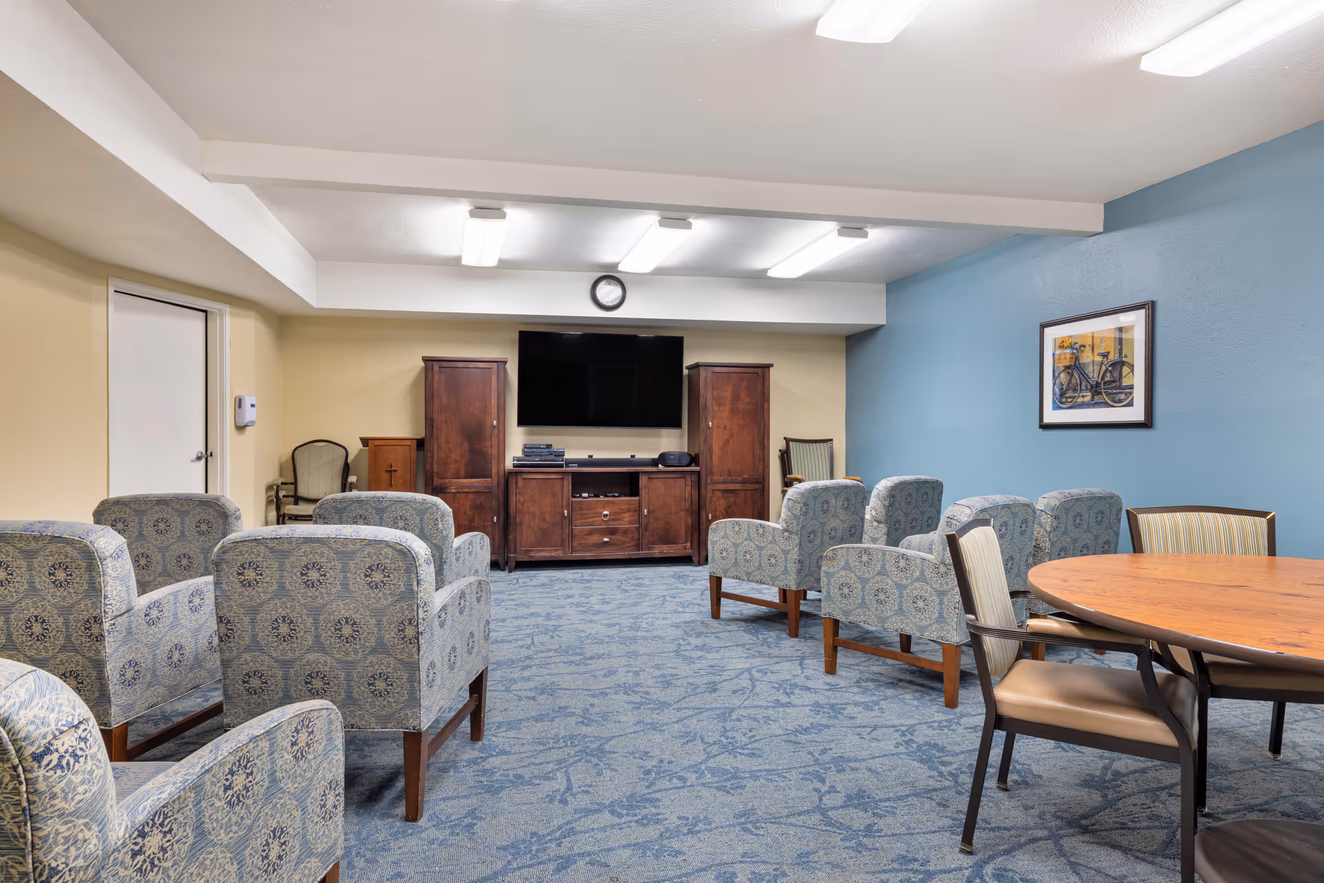 A common area in a senior living facility with multiple patterned armchairs arranged in rows facing a large flat-screen TV mounted on the wall. The room has blue carpet, light yellow and blue walls, wooden cabinets under the TV, a round wooden table with chairs on the right side, and a framed picture of a bicycle on the blue wall.
