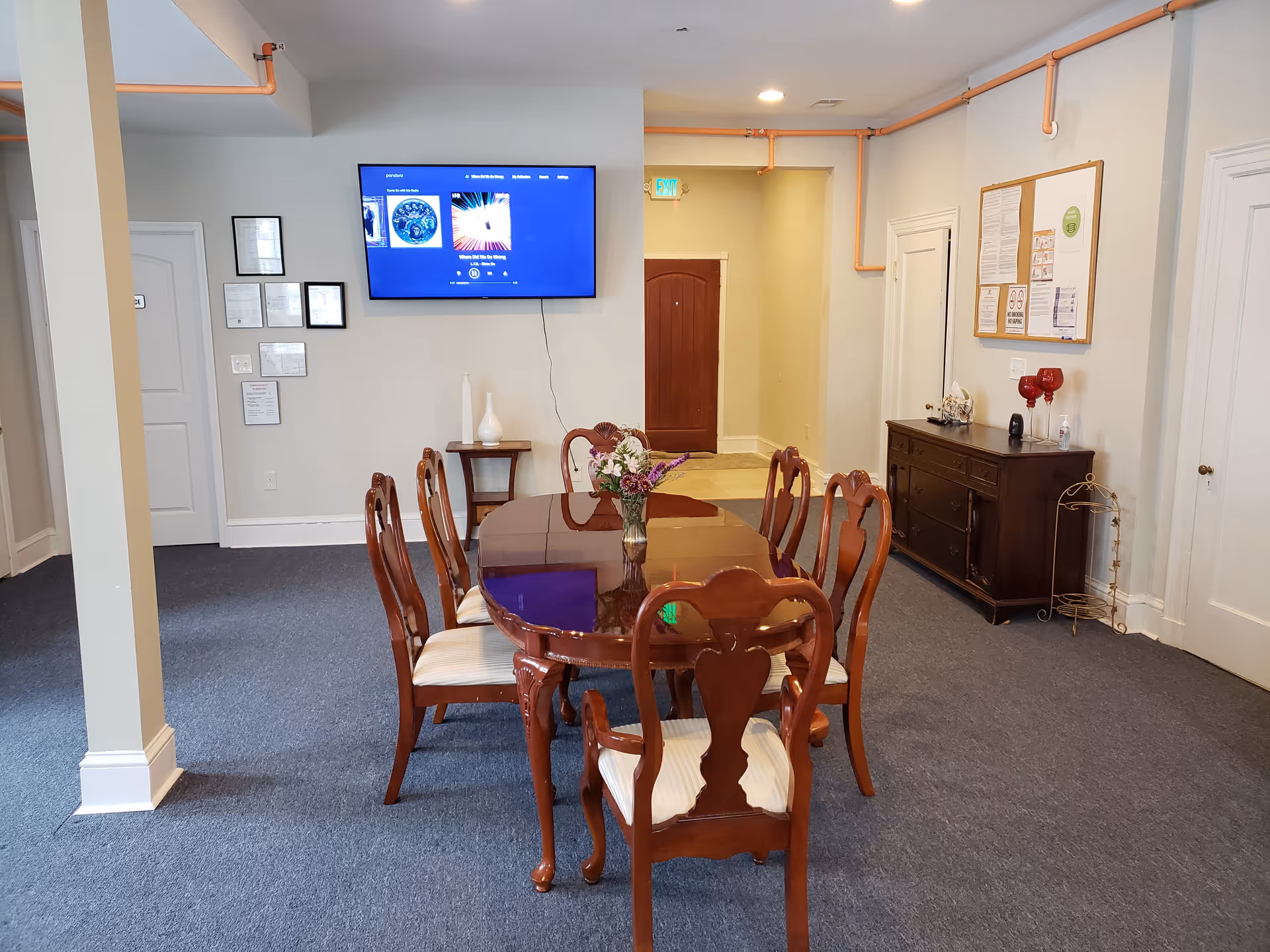 A dining area with a polished wooden table and six matching chairs with white cushions. A vase with flowers is placed on the table. On the wall, there is a flat-screen TV displaying a music player interface. The room has blue carpet flooring, light-colored walls, and some framed documents and a bulletin board on the walls. There is a wooden sideboard with decorative items and a hand sanitizer bottle. The ceiling has exposed orange pipes and recessed lighting.