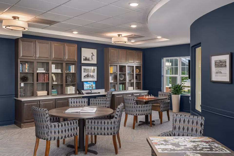 A cozy activity room with dark blue walls and carpeted floor featuring round tables surrounded by patterned chairs. One table has an open book, another has a chessboard, and a third has a partially completed jigsaw puzzle. Built-in wooden cabinets with books and decorative items line the back wall, and framed black-and-white photos hang above them. A large window and a potted plant add natural light to the space.